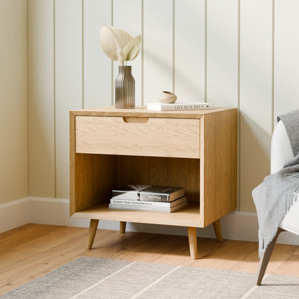 שידת לילה מעץ אלון טבעי עם מגירת אחסון פנימית ומדף פתוח, בעלת רגלי עץ דקיקות ועיצוב מינימליסטי, אידיאלית כחלק מקולקציית שידות לילה לחדר השינה.