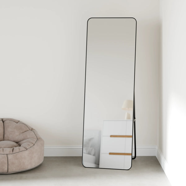 מראה עומדת מלבנית וגבוהה עם מסגרת מתכת שחורה דקה, הנשענת על קיר בחדר שינה מינימליסטי ליד פוף בהיר, משמשת כפריט עיצובי בקולקציית מראות לחדר השינה.